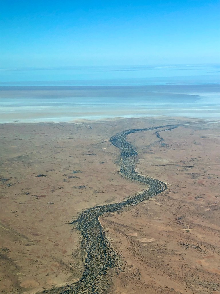 Aerial view of a winding river flowing through a stark desert landscape, with blue sky above and distant salt flats visible.
