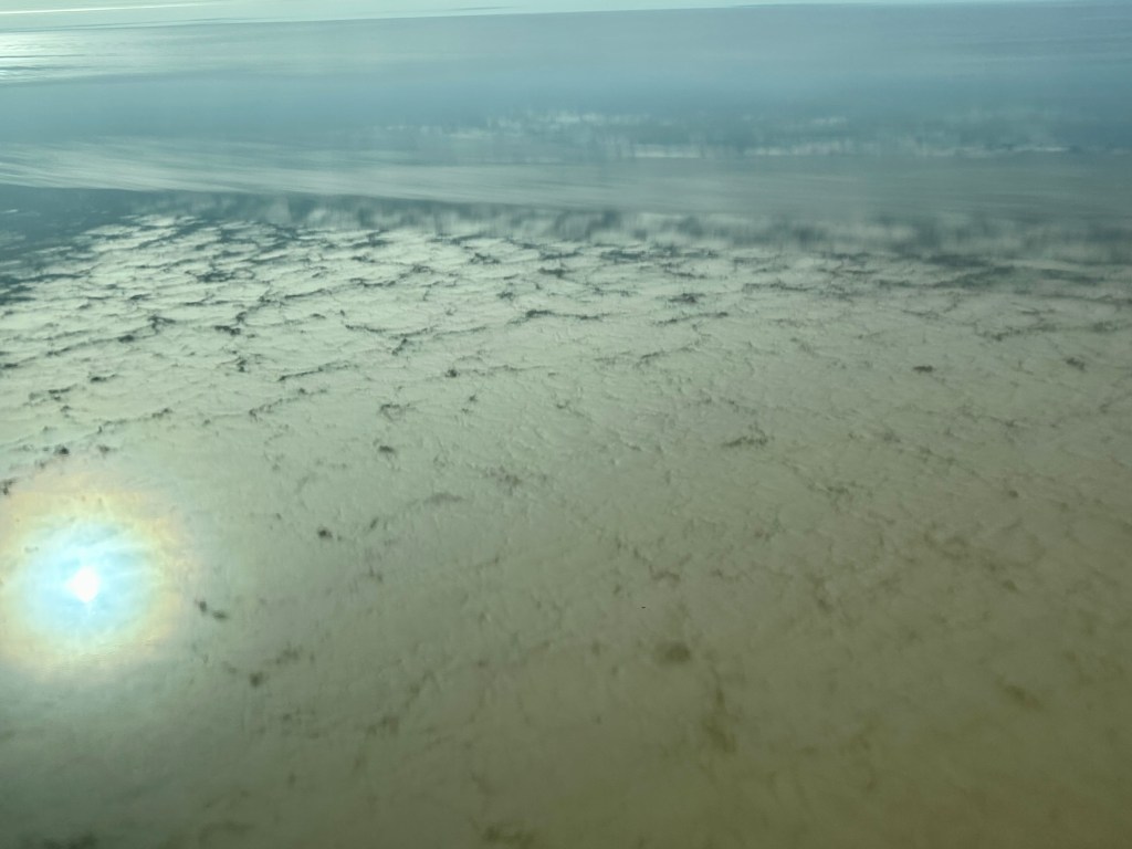 Aerial view of a vast landscape featuring a shimmering body of water reflecting the sun, surrounded by textured land patterns.