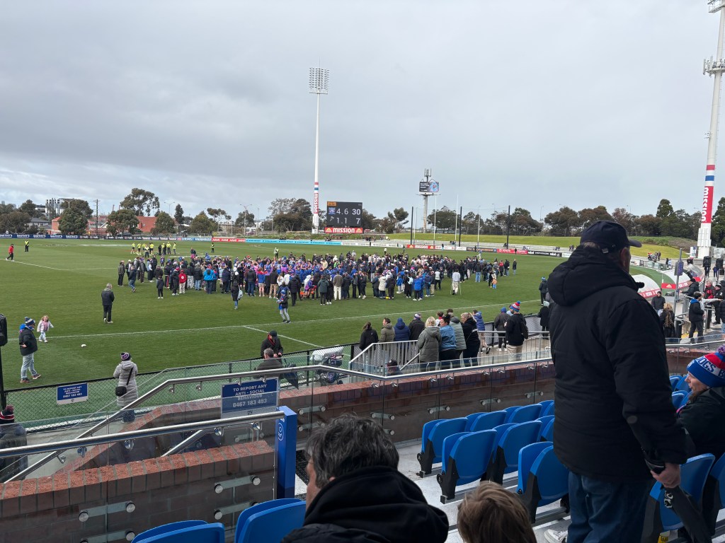 A crowd gathered on the football field after a match, with spectators in the stands and the scoreboard visible in the background.