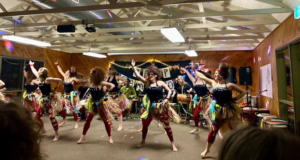 A vibrant performance featuring several dancers dressed in colorful costumes, showcasing traditional dance moves in a lively setting with decorations and musical instruments in the background.