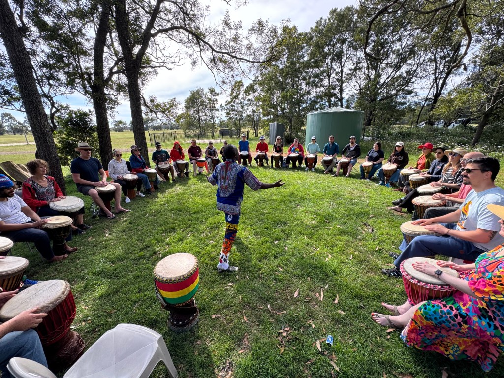 A group of people gathered in a circle, participating in a drumming workshop outdoors, surrounded by trees and green grass.