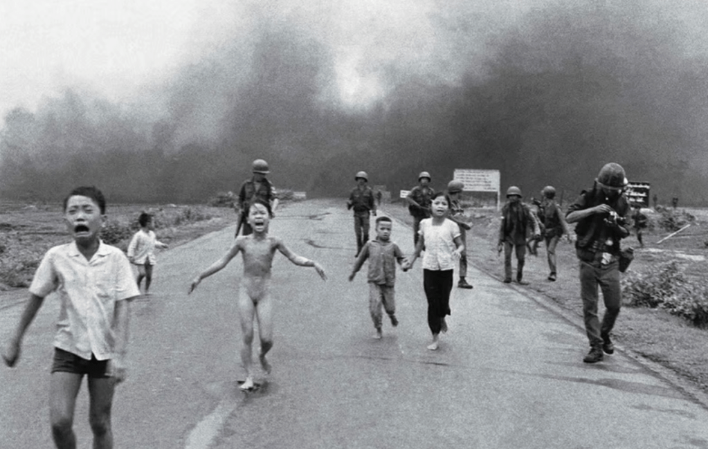 A group of frightened children running away on a road, with soldiers in the background and smoke in the sky, capturing a moment of chaos and fear.