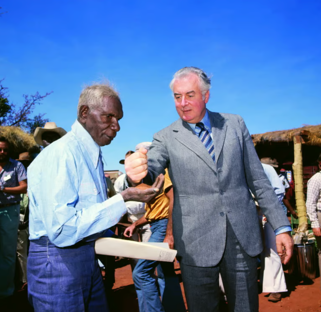 An elderly man in a light blue shirt and trousers extends his hand, while a suited man with gray hair gestures with a clenched fist. They are in a gathering with other people and a clear blue sky in the background.