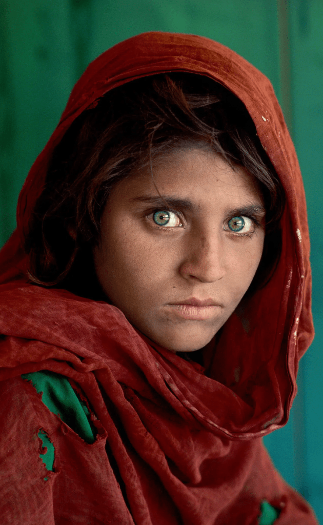 A young girl with striking green eyes and dark hair, wearing a red shawl, looks directly at the camera with a serious expression against a teal background.