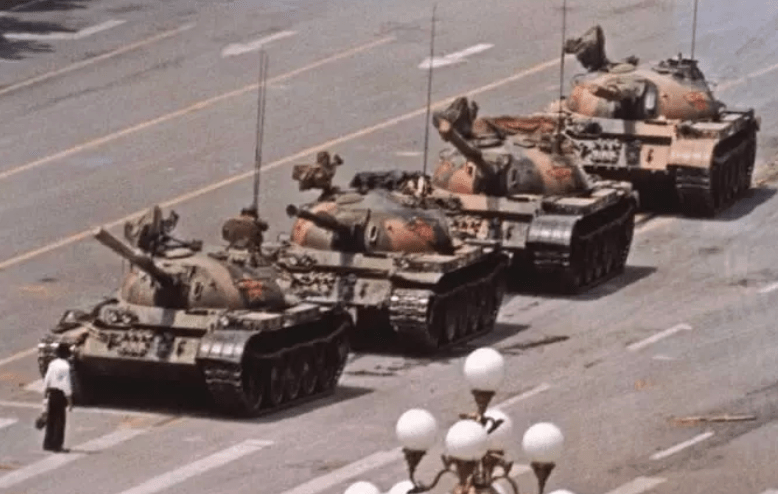 A line of military tanks on a wide avenue, with a lone figure standing in front of them.