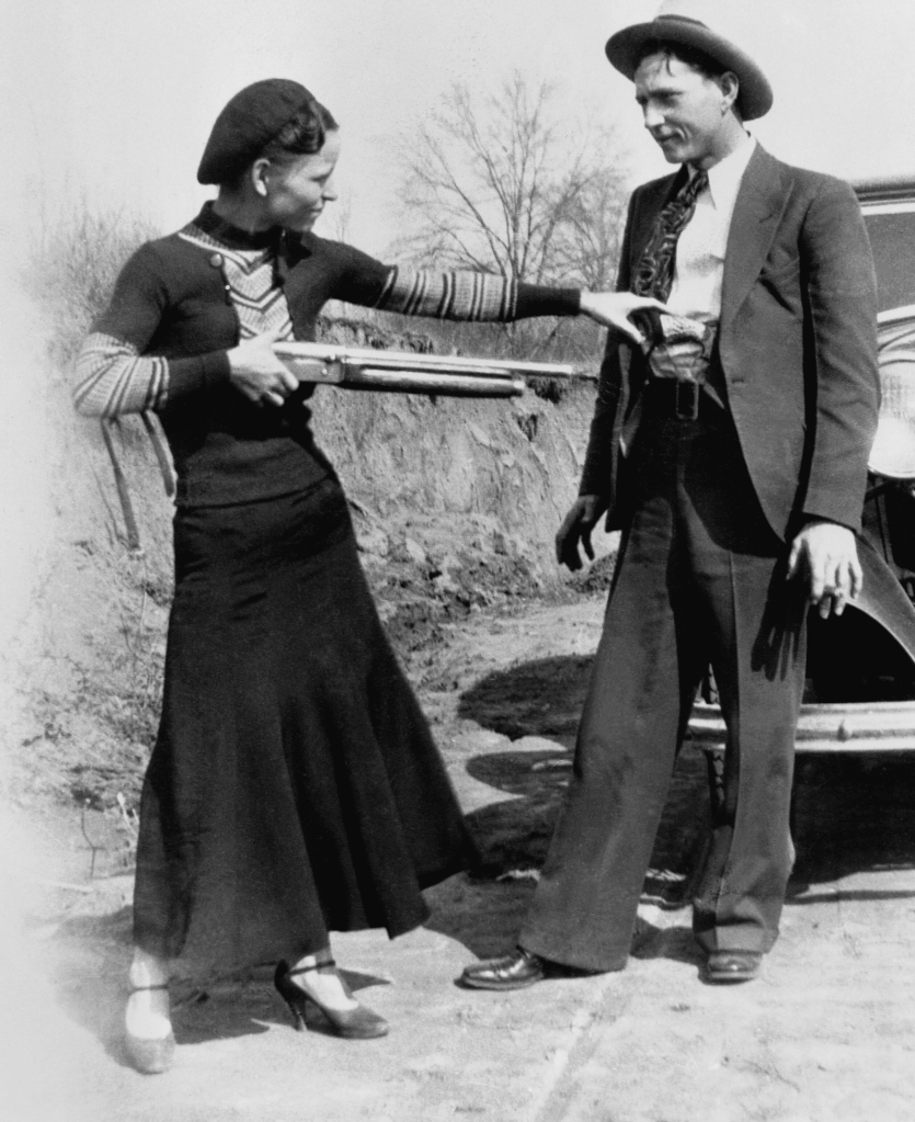 A woman in 1930s attire holds a shotgun while pointing it at a man dressed in a suit, who appears surprised. They are standing outdoors near an old car, with trees in the background.