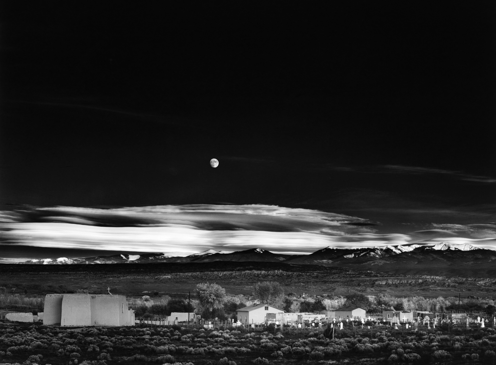 Black and white landscape featuring a village with adobe structures, set against a backdrop of mountains and a partially clouded sky, with a full moon visible.