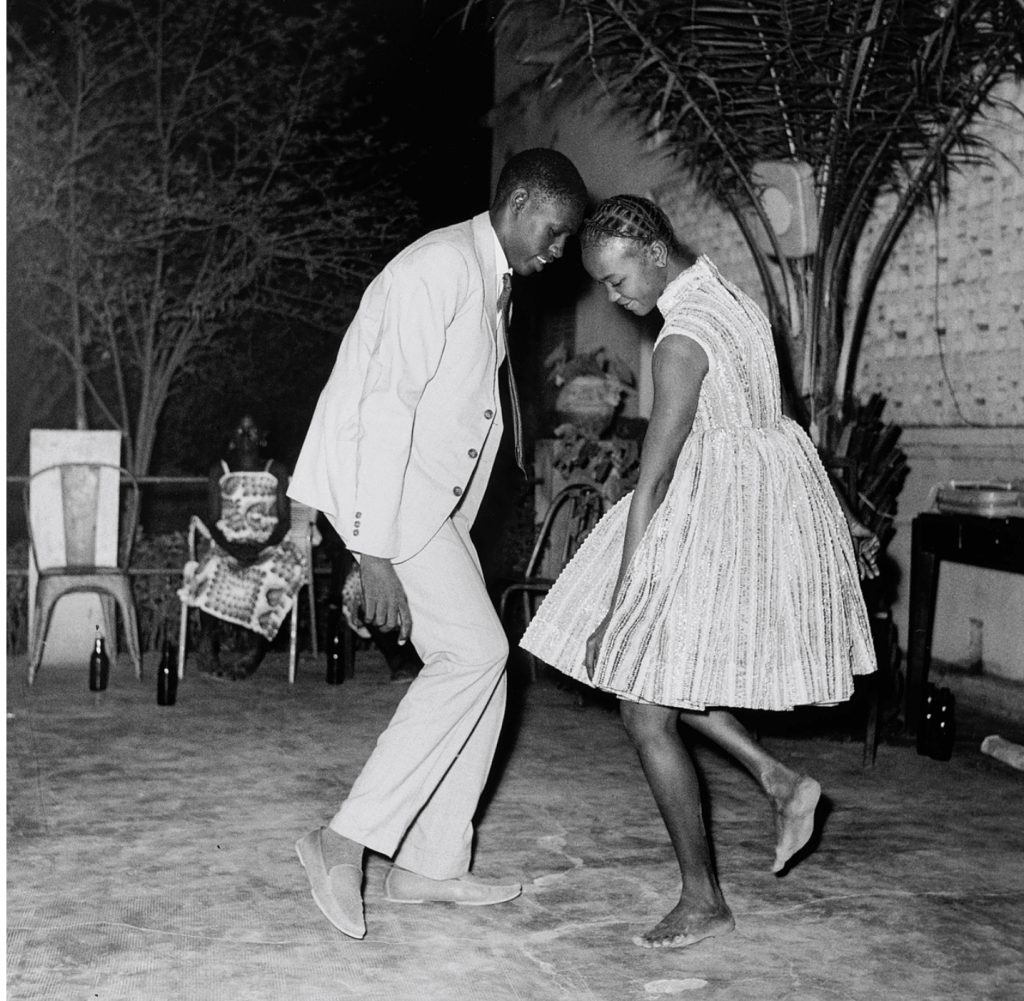 A black and white photo of a man and woman dancing closely together, with the woman wearing a short, shiny dress and the man in a light suit. In the background, there are chairs and a palm tree, and a seated figure can be seen.