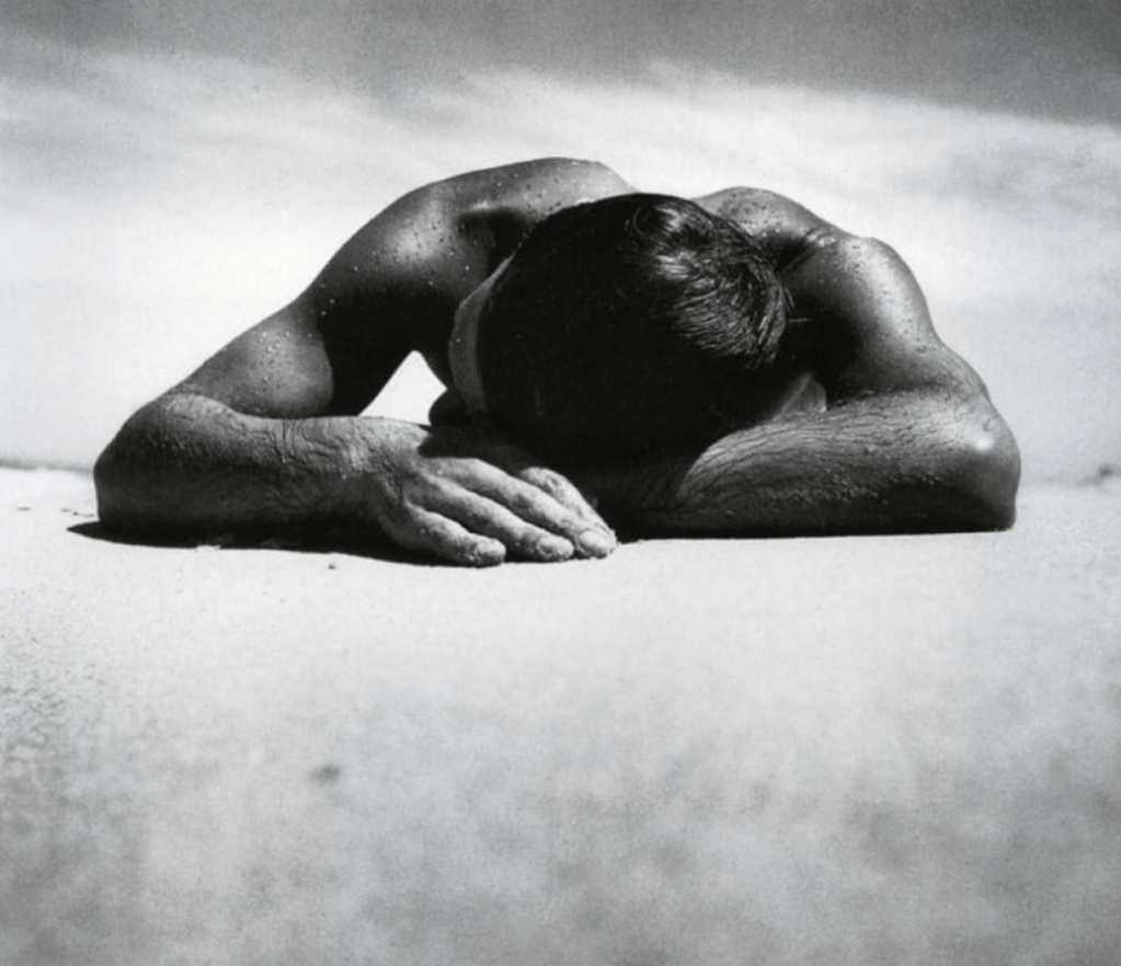 A black and white photograph of a person lying face down on a sandy surface, with their arms crossed under their head.
