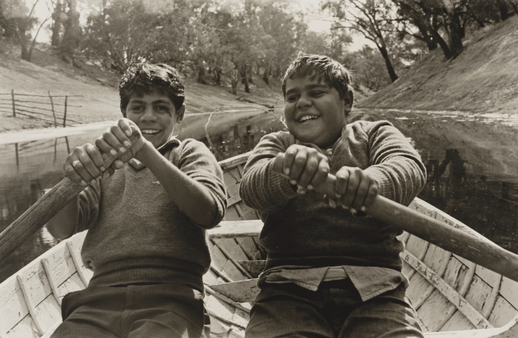Two young boys rowing a boat together on a calm river, smiling and enjoying their time outdoors.