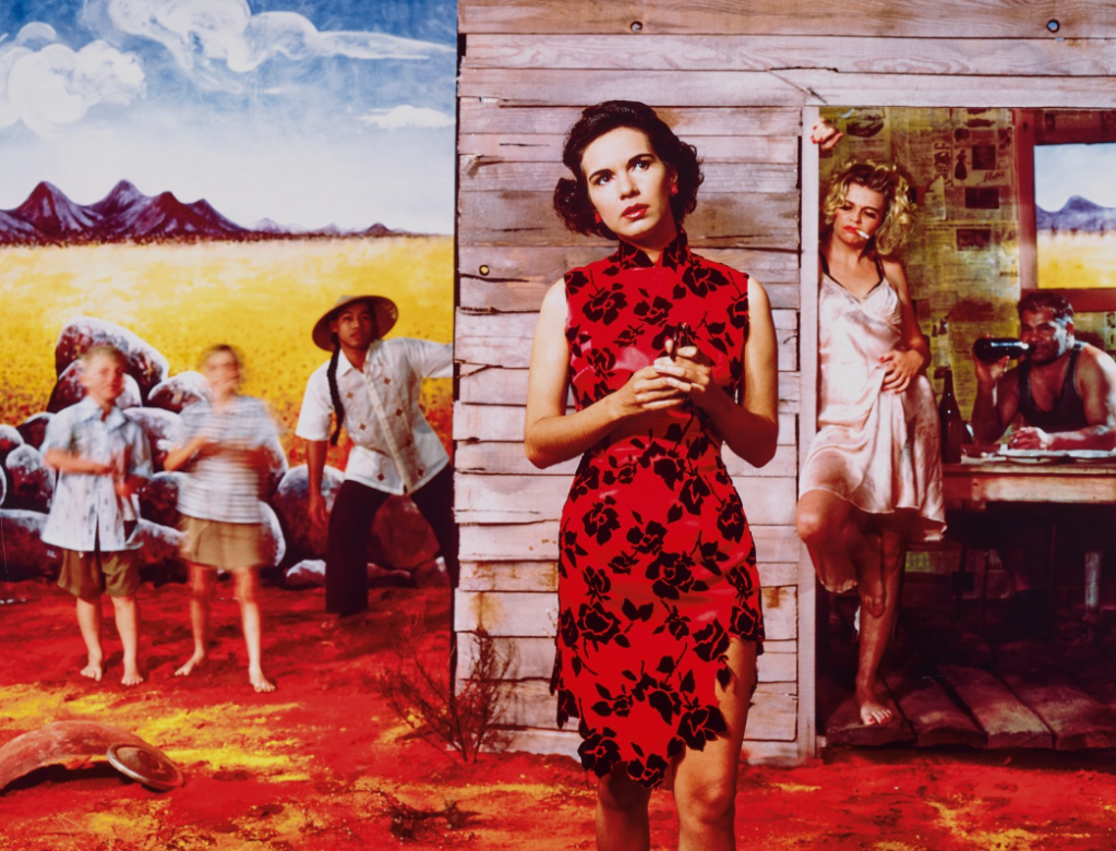 A woman in a red floral dress stands in front of a wooden shed, holding a small object, while another woman in a light dress leans against the door. Two children and a man in a hat are visible in the background, with a vibrant landscape consisting of yellow fields and mountains.