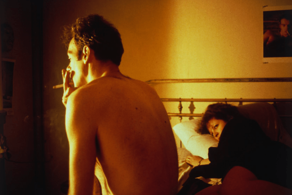 A shirtless man smoking a cigarette in a dimly lit room, with a woman lying on a bed in the foreground, looking contemplative.