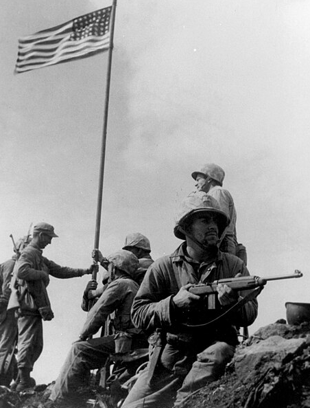A group of soldiers raising the American flag on a hill during a military operation, with one soldier holding a rifle and others assisting in the flag raising.