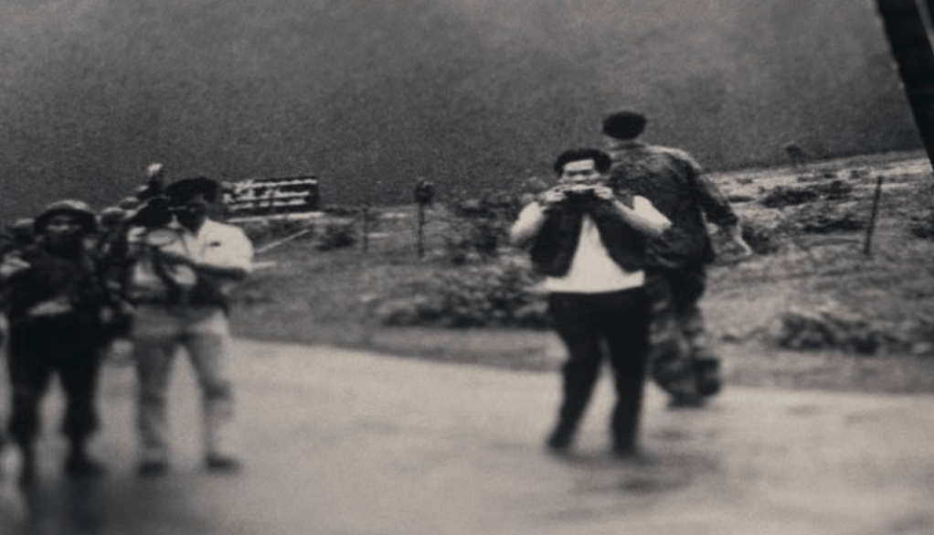 A historical black and white image showing a group of individuals on a rural road. One person in the foreground is holding a camera and facing the viewer, while another person next to him is operating a film camera. In the background, a soldier is walking away, and a sign is partially visible.