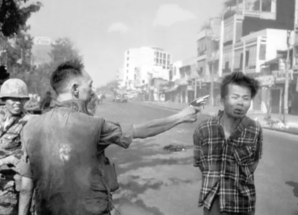 A soldier aims a gun at a bound man on a street, with a second soldier visible in the background. The scene appears tense and somber, reflecting a moment of conflict.