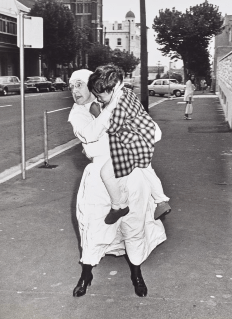 A woman in a headscarf carries a child on her back, both looking into the distance. They are on a sidewalk with trees and buildings in the background. Other people and parked cars are also visible.