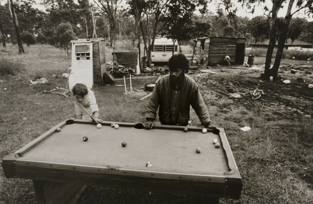 A young child and an adult man playing pool outdoors at a rundown table, surrounded by an unkempt yard with dilapidated structures in the background.