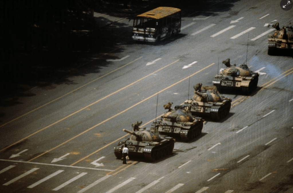 A lone figure stands in front of a row of military tanks on a wide street, with a bus in the background.