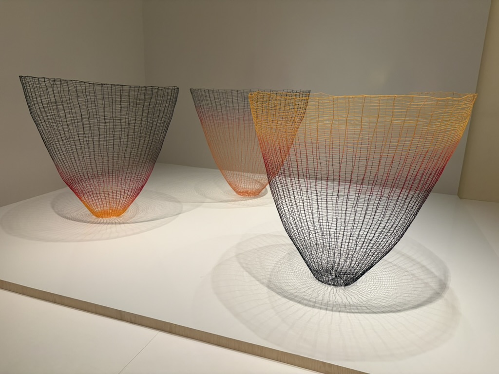 Three large, colorful wire sculptures with a conical shape, featuring gradients of orange, red, and black, set on a white display surface.