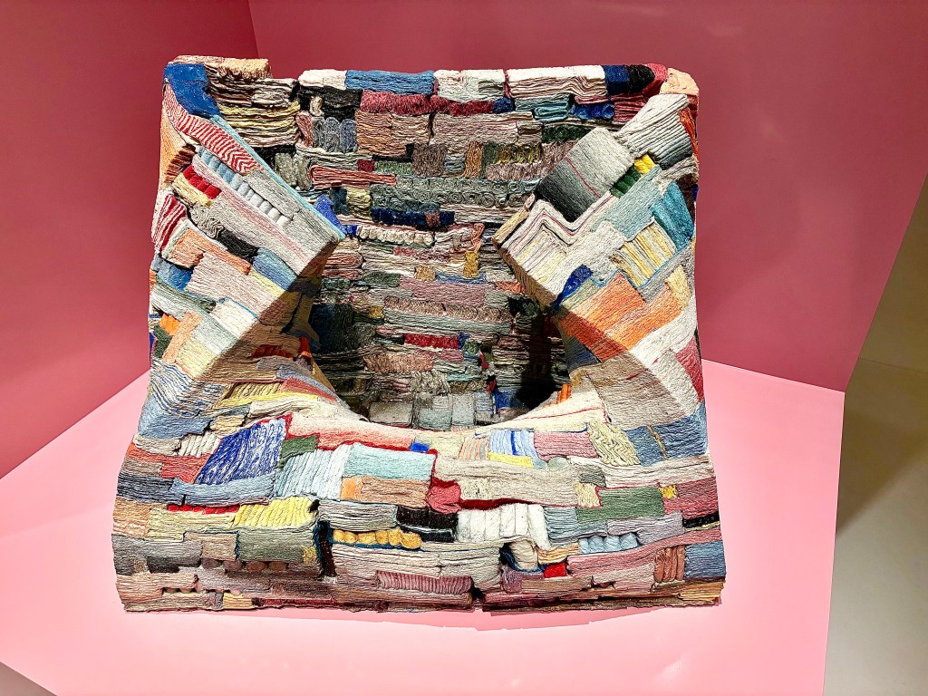 A colorful, textured sculpture resembling a seat, made from layered fabric strips, displayed against a pink background.
