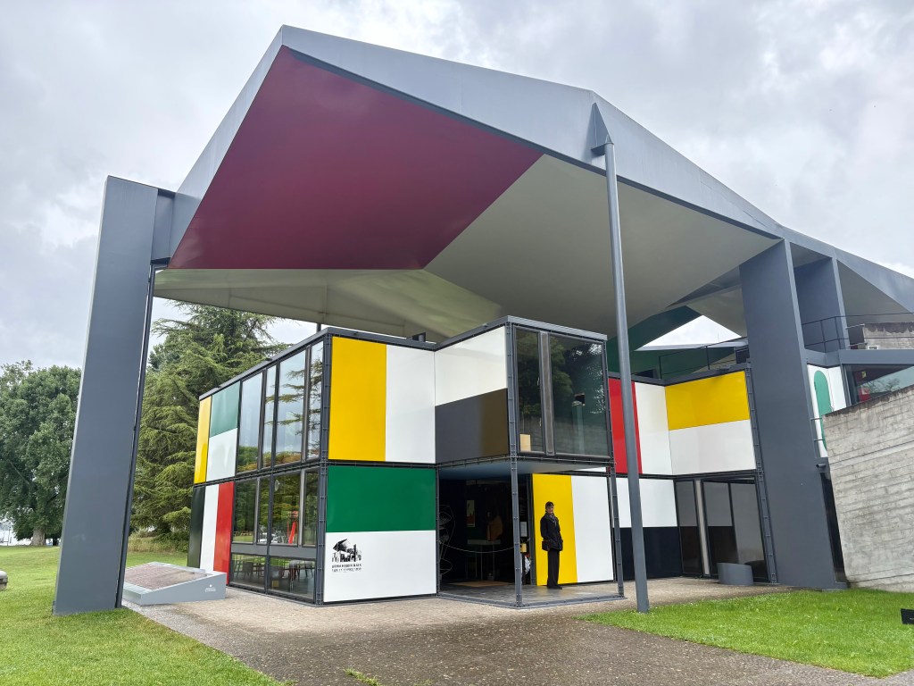 A modern architectural building featuring a colorful facade with large windows and a distinctive polygonal roof, surrounded by green grass and trees.