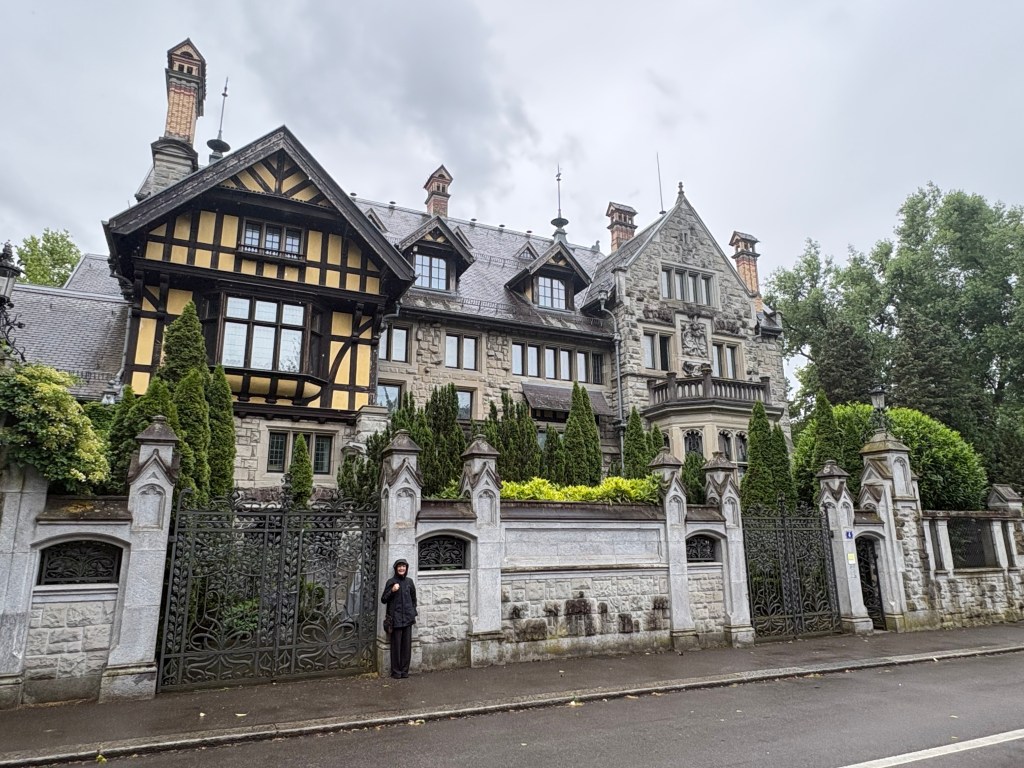 A large, historic mansion with a combination of stone and wood architecture, featuring intricate details and a manicured garden, with a person standing near the entrance gate.