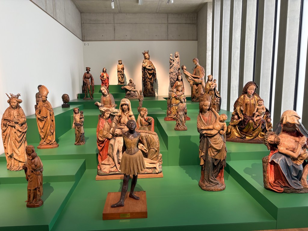 A gallery displaying a variety of wooden sculptures, including religious figures and historical characters, arranged on green platforms under bright lighting.