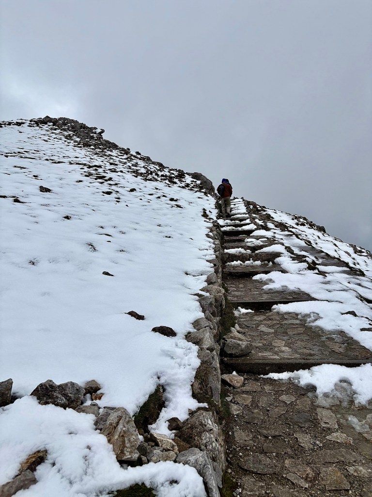 A hiker ascends a snow-covered mountain trail, with rocky slopes and overcast skies above.