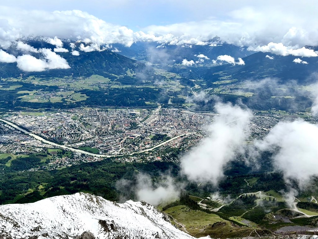 Aerial view of a town nestled in a valley surrounded by mountains, with lush greenery and patches of clouds in the sky.