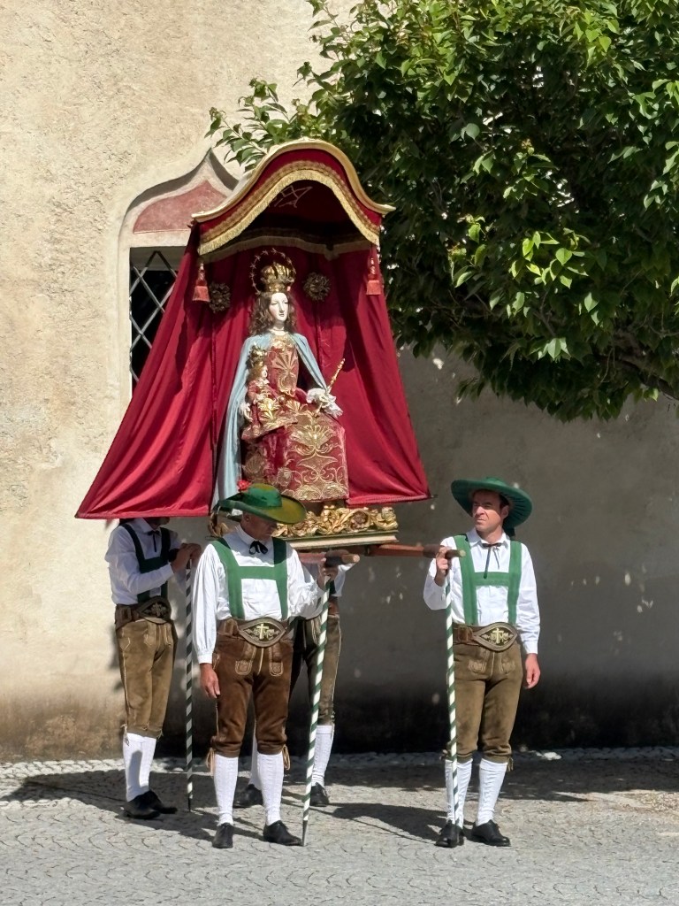 A group of four men in traditional attire carrying a large, decorated canopy with a statue of a seated figure underneath, surrounded by greenery and a textured wall.