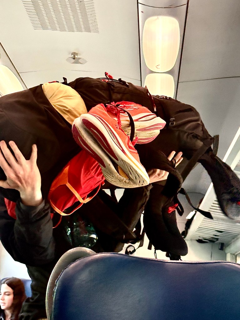 A person struggling to carry an oversized backpack filled with various items, including running shoes and clothing, while seated in a train.