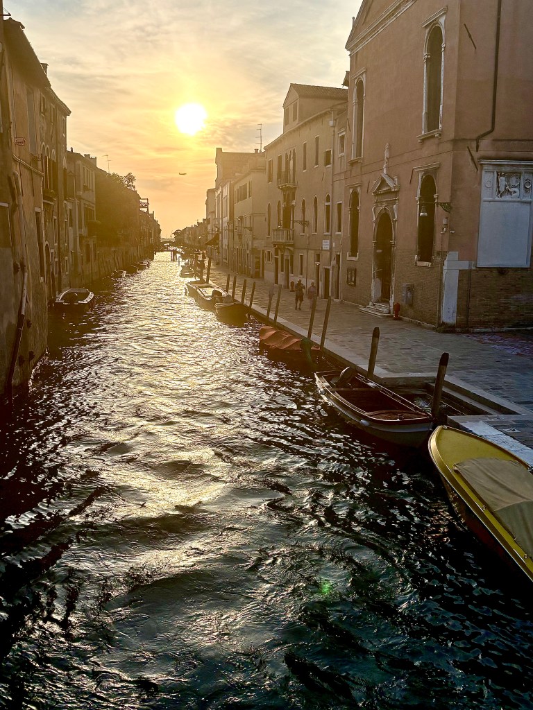 A picturesque canal in a city at sunset, featuring calm water reflecting the orange and yellow hues of the sky, with gondolas lined along the edges and historic buildings on either side.