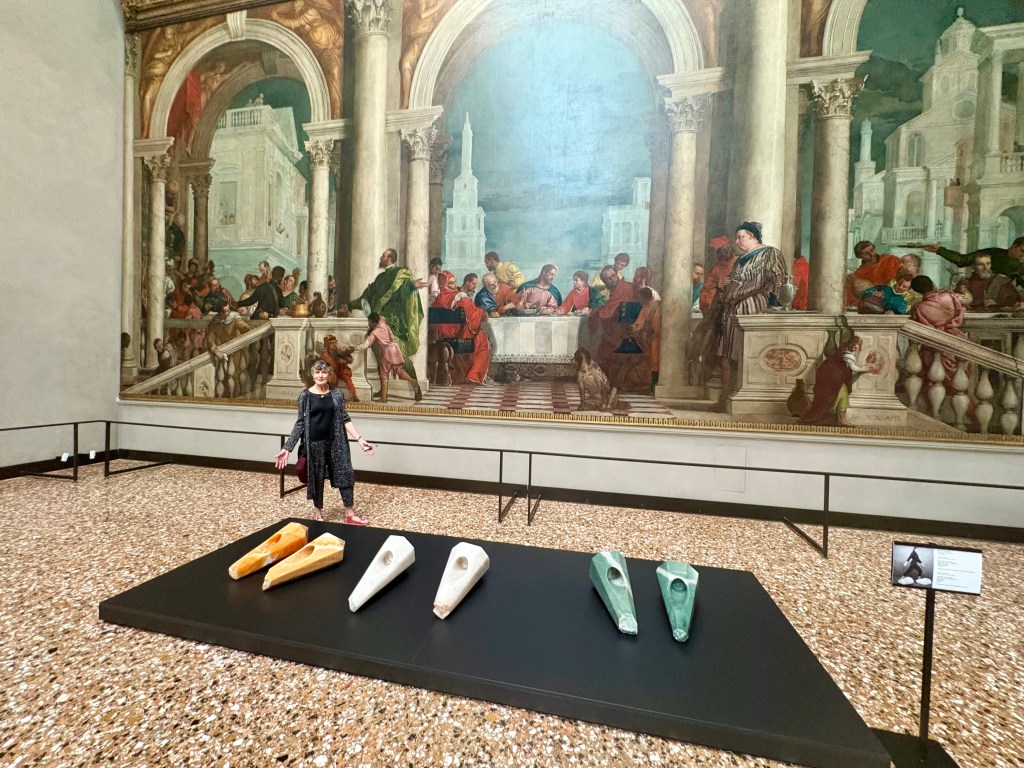 A woman stands in an art gallery in front of a large historical mural depicting a gathering, alongside a display of five abstract sculptural pieces on a black platform.