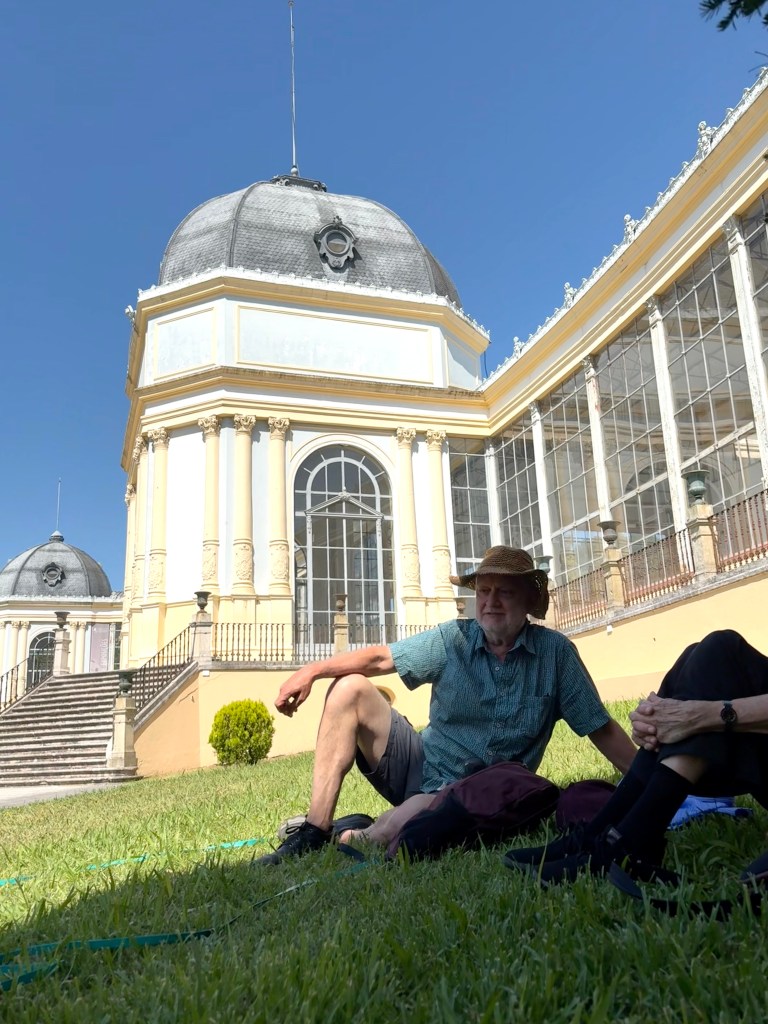 A man in a straw hat sitting on grass in front of an ornate building with large glass windows and a domed roof on a sunny day.