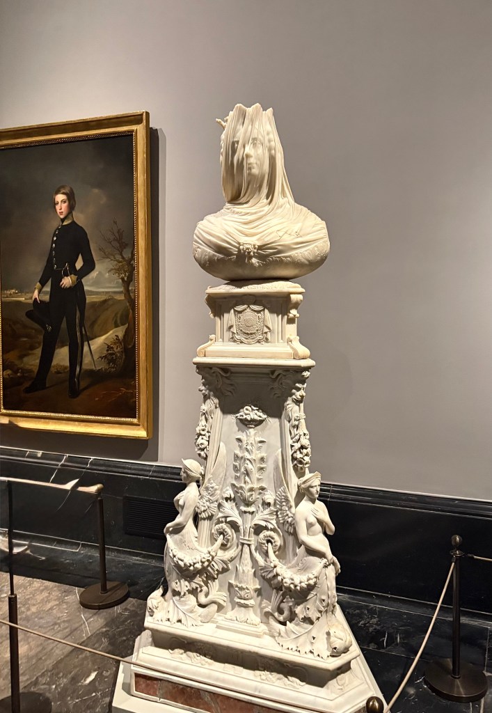 A white marble bust draped in a veil and resting on an ornate pedestal, featuring intricate carvings and figures, displayed in an art gallery next to a painting of a young man in a military uniform.