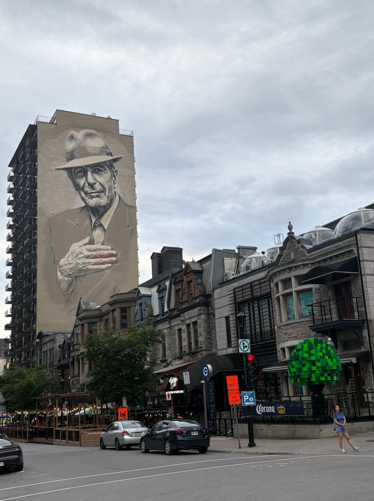 A large mural of an elderly man in a hat on a building, with a city street and vintage architecture in the foreground, featuring shops and trees.