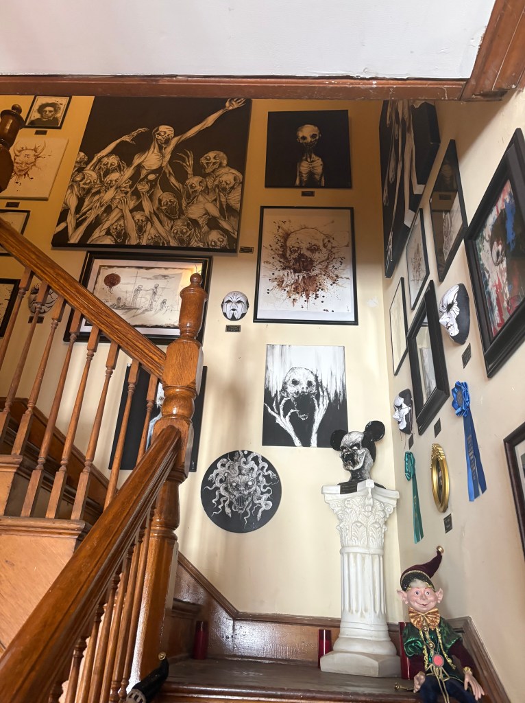 A staircase leading up with walls adorned with various dark-themed artworks, including sketches of skeletal figures and eerie masks, alongside a decorative statue and a whimsical doll at the base.