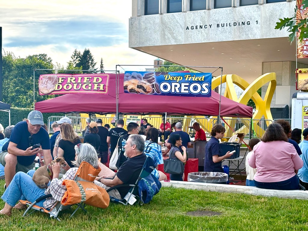 A busy outdoor festival scene with a tent offering fried dough and deep-fried Oreos, surrounded by people enjoying the event, some seated on the grass.