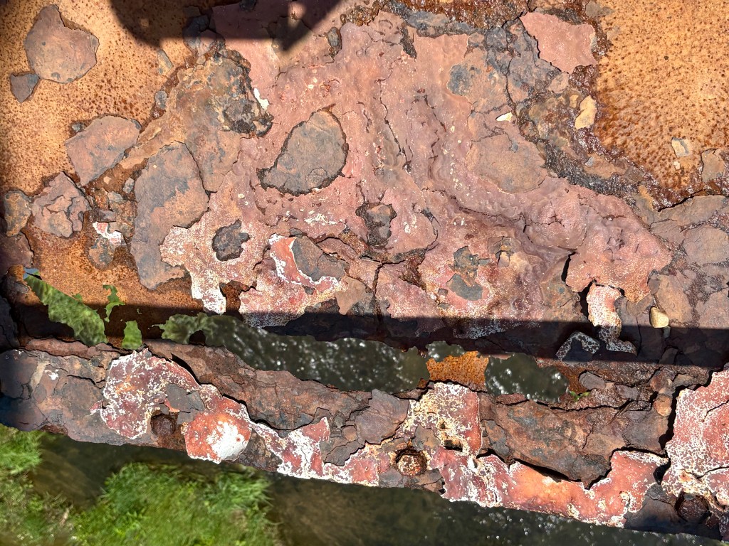 Close-up view of rusty, corroded metal surface with peeling paint, revealing a textured orange and brown surface and a glimpse of water below.