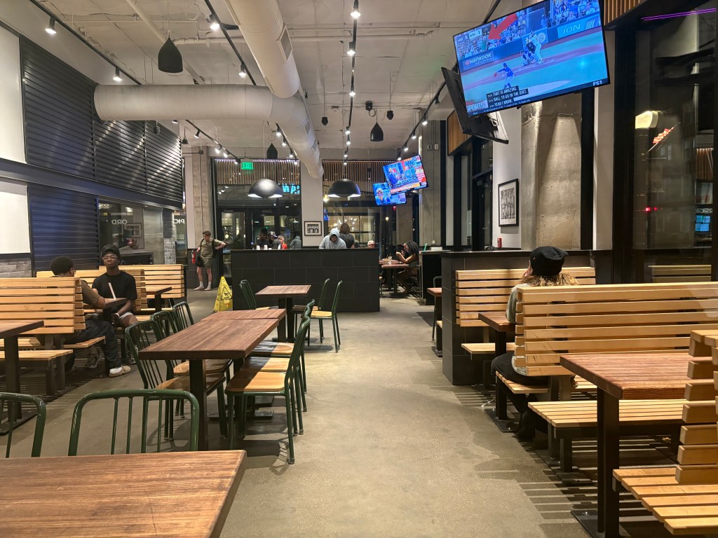 Interior of a modern restaurant featuring wooden tables and benches, with several patrons seated, and multiple televisions displaying sports.