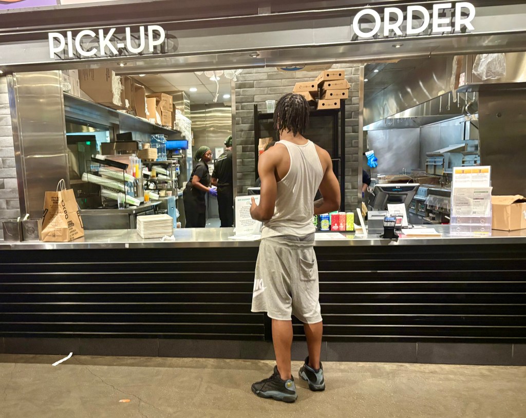 A person wearing a tank top and shorts stands in front of a food pickup counter, waiting to collect their order. Staff members can be seen in the background preparing food.