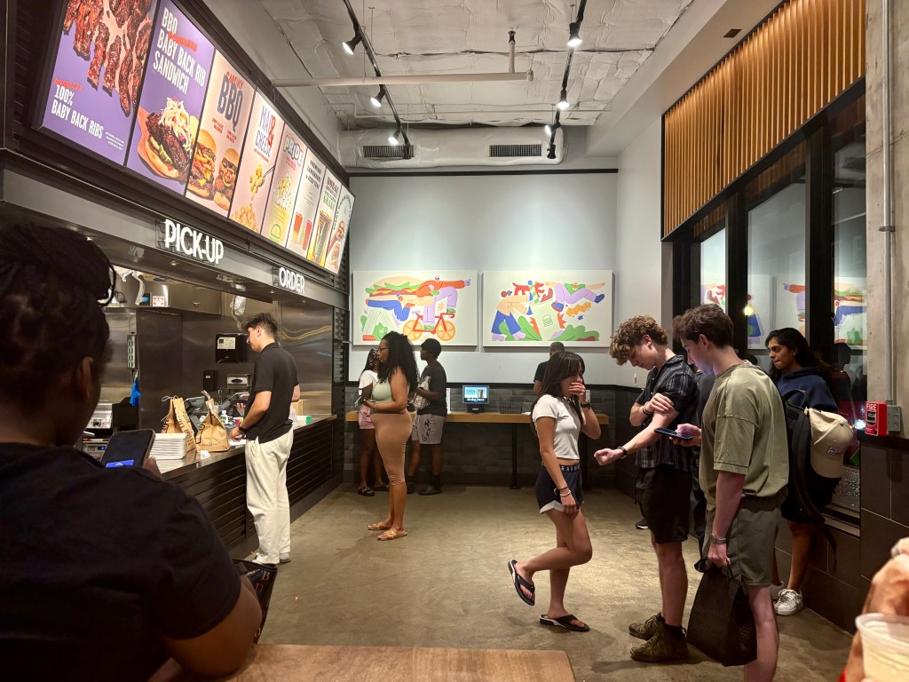A modern restaurant interior featuring a counter with a menu displaying BBQ items. Customers are lined up to order, and some are engaged in conversation. Colorful artwork adorns the walls.
