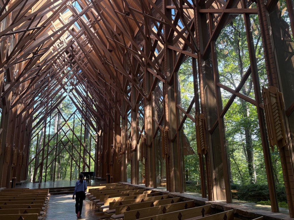 Interior of a modern wooden chapel with a high, angular ceiling and large windows, surrounded by trees, featuring a person walking down the aisle.