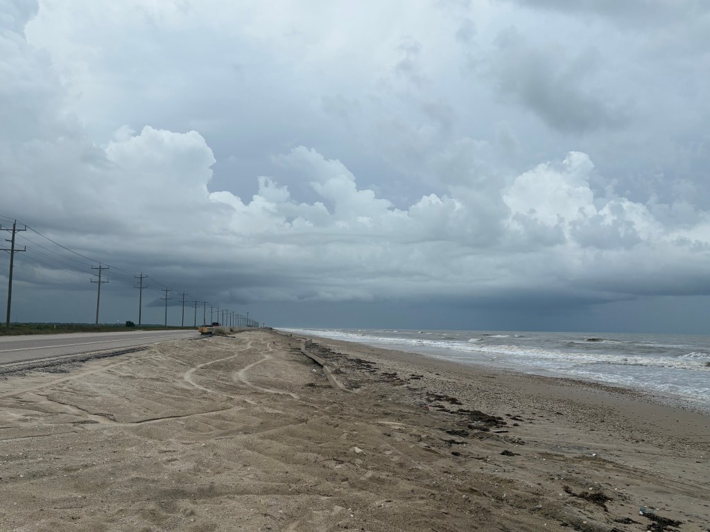 Coastal view showing a sandy beach, ocean waves, and cloudy gray sky, with power lines and a road in the distance.