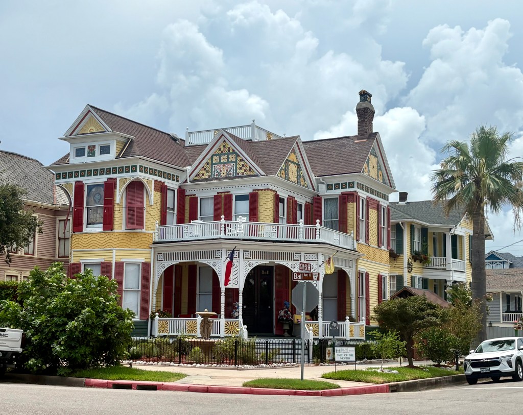 Colorful Victorian-style house with yellow siding and red shutters, featuring a large porch and decorative cornices, set against a cloudy sky.