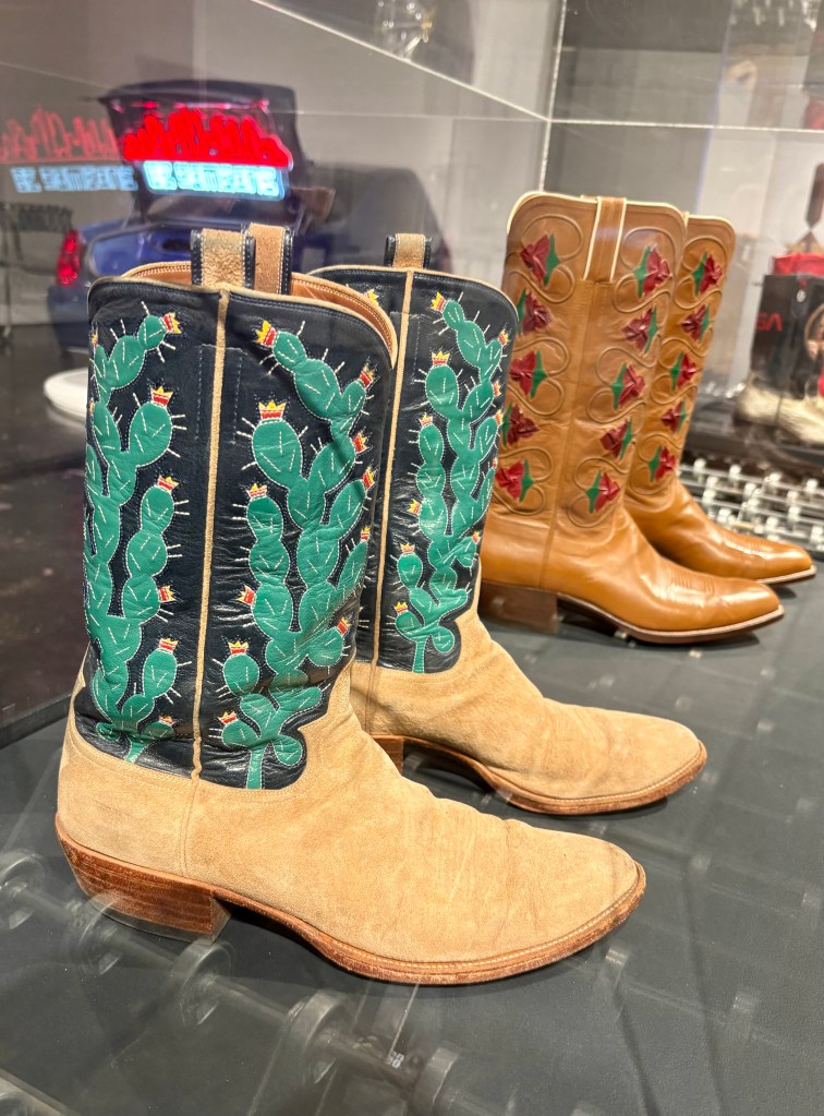 A pair of intricately designed western cowboy boots featuring green cactus embroidery on a black background, with a tan pair of boots displayed beside them.