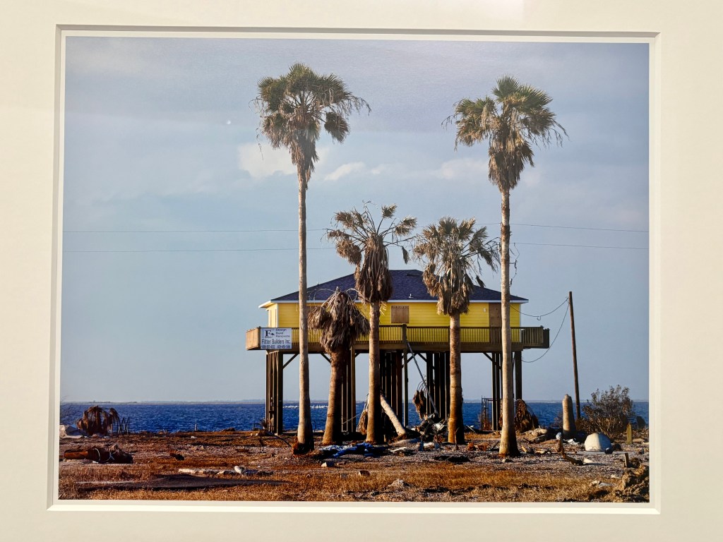 A raised yellow house supported by stilts, surrounded by palm trees, with a blue ocean in the background and a clear sky.
