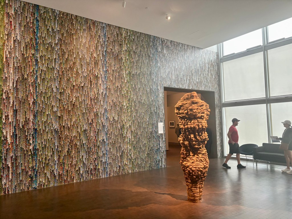 An art gallery featuring a large, textured sculpture in the foreground and a colorful abstract wall installation in the background. Two visitors are walking through the space.