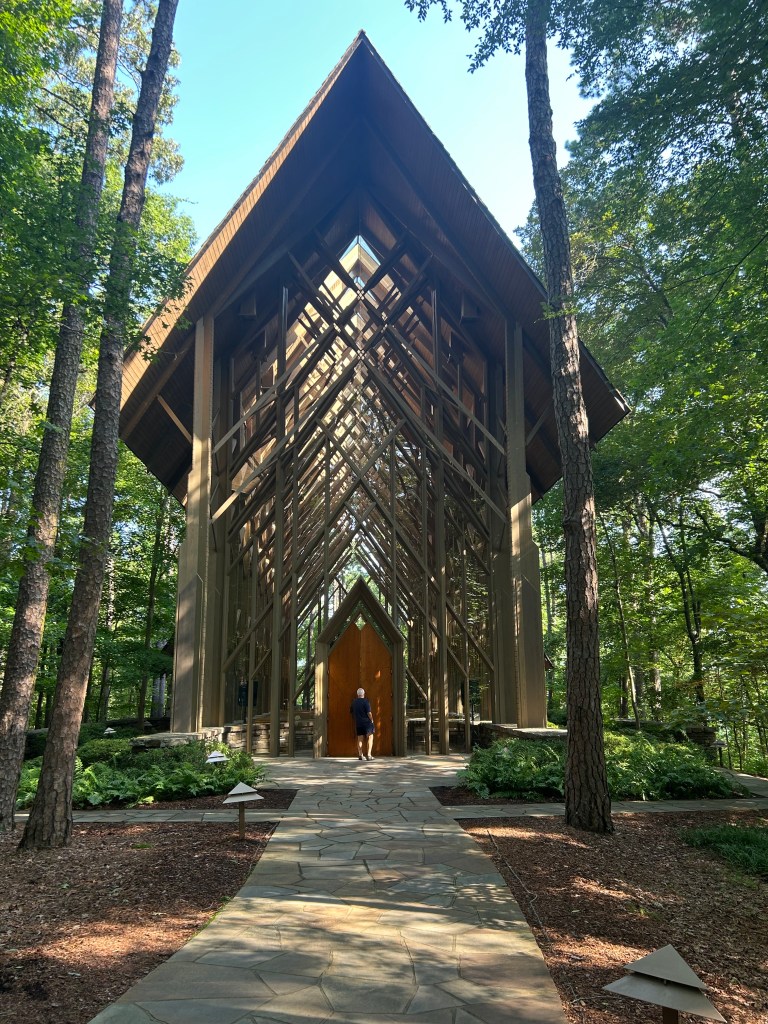 A person walking towards a modern, architecturally striking wooden chapel surrounded by trees and greenery.