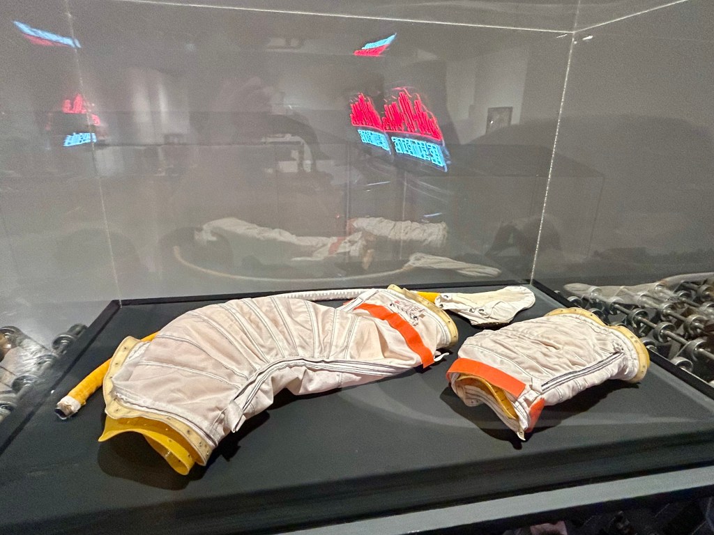 A display case containing a pair of vintage white and orange astronaut gloves, showcasing intricate stitching and details, with a reflective background.